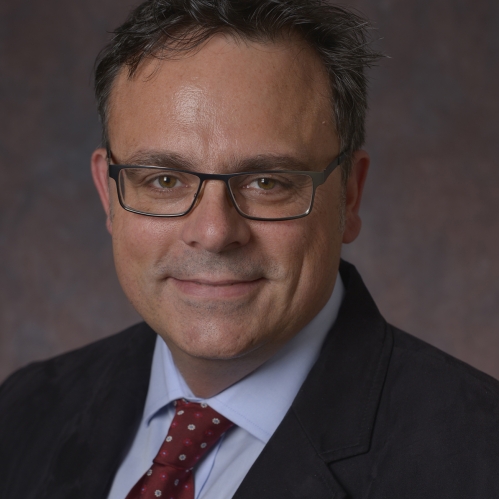 head shot of male with eyeglasses and short dark brown hair wearing a dark blue suit, light blue shirt, and maroon pattern tie