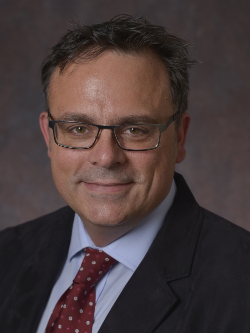 head shot of male with eyeglasses and short dark brown hair wearing a dark blue suit, light blue shirt, and maroon pattern tie