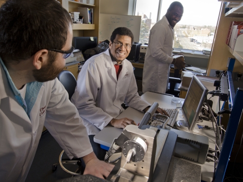 Three men in white lab coats interacting with each other as they conduct research in a biomedical engineering lab. 