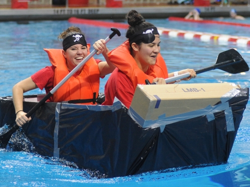 Two female students wearing eye patches and oragne life vests participate in cardoard canoe races in an indoor pool.