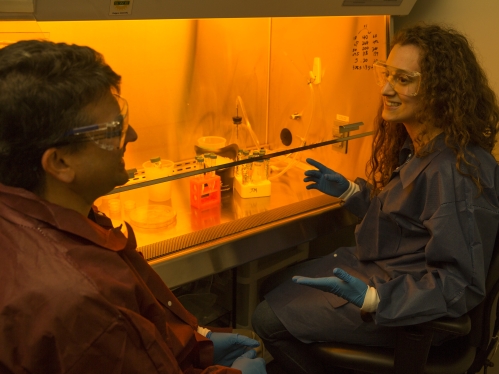 White female student with brown curly hair wearing safety googles, blue lab coat and blue gloves speaks to a white male professor also wearing safety glasses. They are sitting in front of a hooded lab bay that is lit with a yellow light.