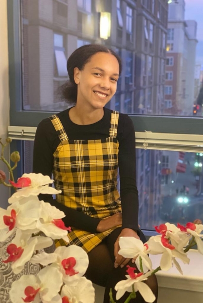 Young Black woman sits on a window sill with a city view behind her and white and red flowers in the foreground. She is wearing a yellow and black plaid jumper with a black shirt.