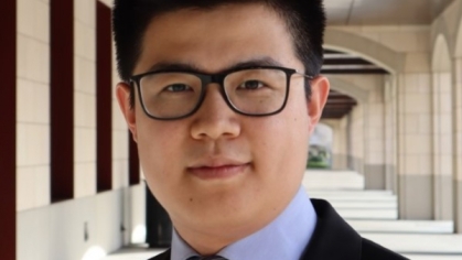 head shot of asian man with short black hair wearing dark rimmed eyeglasses, a black suit, light blue shirt, and blue tie