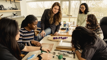 Five female students working on project with the assistance of a female professor.