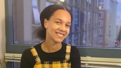 Young Black woman sits on a window sill with a city view behind her and white and red flowers in the foreground. She is wearing a yellow and black plaid jumper with a black shirt.