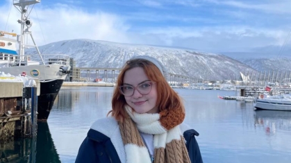 Young woman with red hair wearing a heavy coat and brown striped scarf stands near a harbor in Norway with fishing boats and mountains in the background.