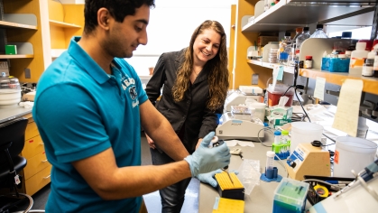 Female professor in a science lab instructing a male student who is using a syringe to test material.