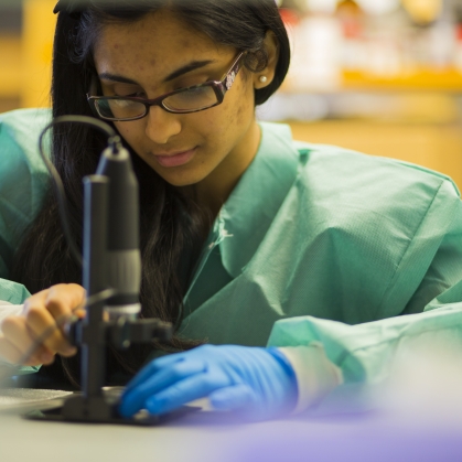 Female student wearing safety glasses, a green lab coat, and one blue latex glove as she works with a microspace.