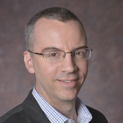 head shot of man with short hair wearing eyeglasses and wearing a dark grey suit with white, blue checkered shirt