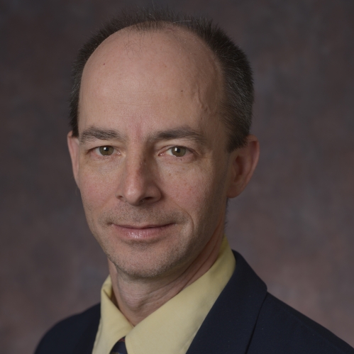 head shot of male wearing a black suit, yellow shirt, and dark patterned tie