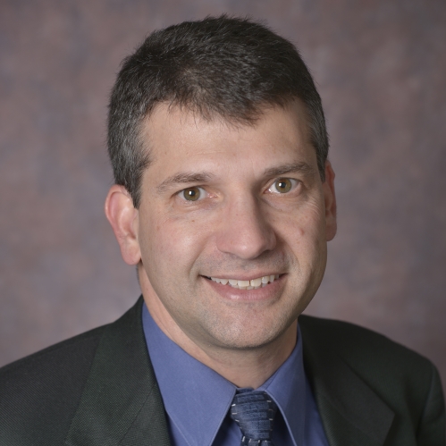 head shot of man with short brown hair wearing a dark grey suit, blue shirt and greyish blue patterned tie