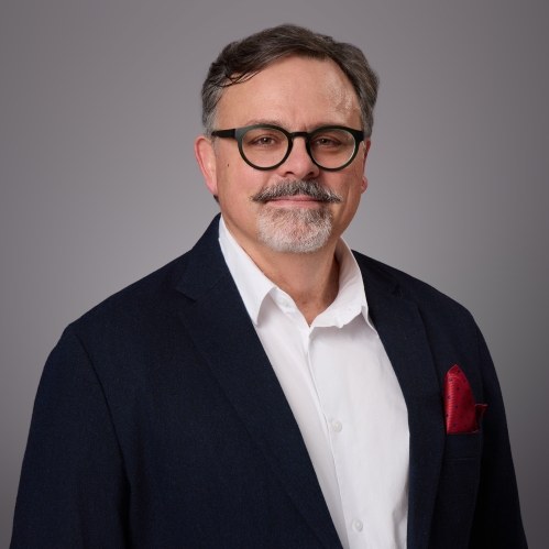Headshot of male with short hair, grey beard and moustache, and eyeglasses wearing a black suit jacket, white button down shirt, and a red lapel handkerchief.