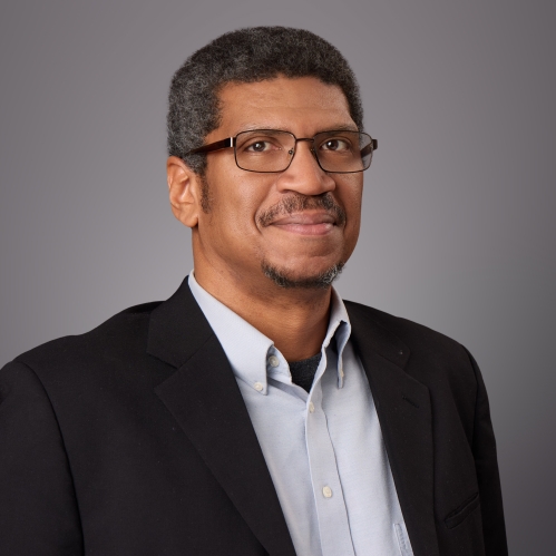 Headshot of Black male with eyeglasses wearing a light grey button down shirt and a dark suit jacket.