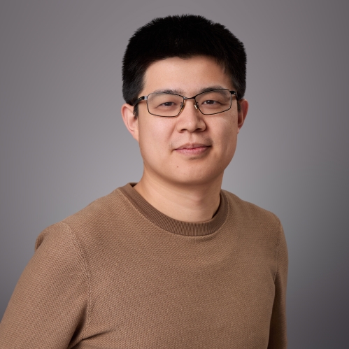 Headshot of male with short black hair and eyeglasses wearing a brown shirt.