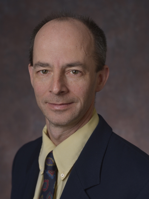 head shot of male wearing a black suit, yellow shirt, and dark patterned tie