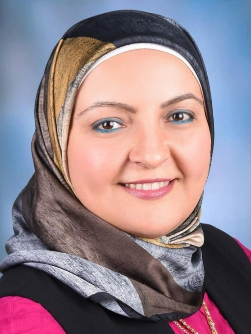 Female wearing a grey and brown head scarf, and a pink and black shirt with a necklace.