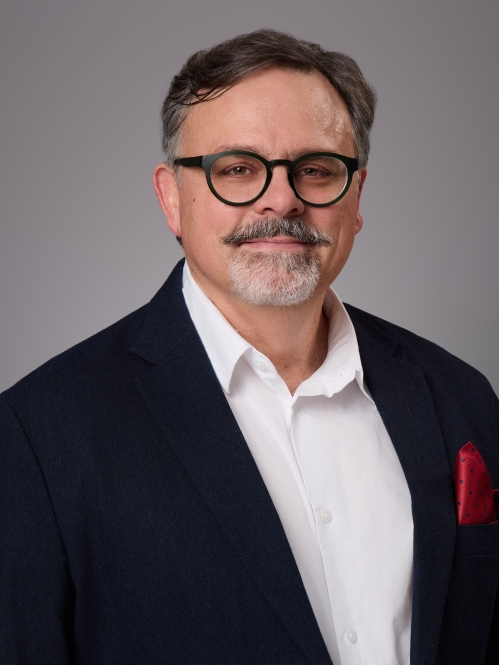 Headshot of male with short hair, grey beard and moustache, and eyeglasses wearing a black suit jacket, white button down shirt, and a red lapel handkerchief.