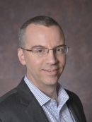 head shot of man with short hair wearing eyeglasses and wearing a dark grey suit with white, blue checkered shirt