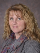 head shot of woman with long blonde curly hair wearing a grey sweater with a grey scarf draped aroung her neckaround