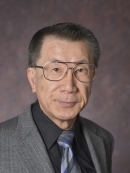 head shot of man with short dark grey hair and eyeglasses wearing a grey suit with a dark grey shirt, and blue, grey striped tie