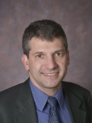 head shot of man with short brown hair wearing a dark grey suit, blue shirt and greyish blue patterned tie