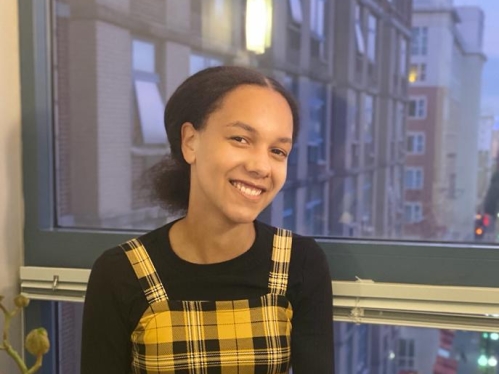 Young Black woman sits on a window sill with a city view behind her and white and red flowers in the foreground. She is wearing a yellow and black plaid jumper with a black shirt.