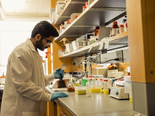 Male student wearing safety glasses, a white lab coat, and blue latex gloves working with a pipette in a lab.