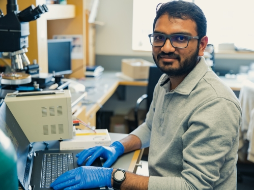 Male student with beard and glasses working at a computer in a lab.