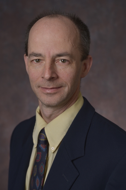 head shot of male wearing a black suit, yellow shirt, and dark patterned tie