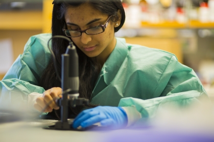 Female student wearing safety glasses, a green lab coat, and one blue latex glove as she works with a microspace.