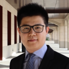 head shot of asian man with short black hair wearing dark rimmed eyeglasses, a black suit, light blue shirt, and blue tie